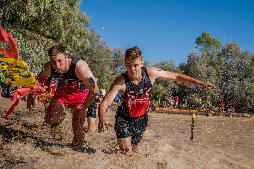 Two men in singlets struggle for the lead of a race through brown water 