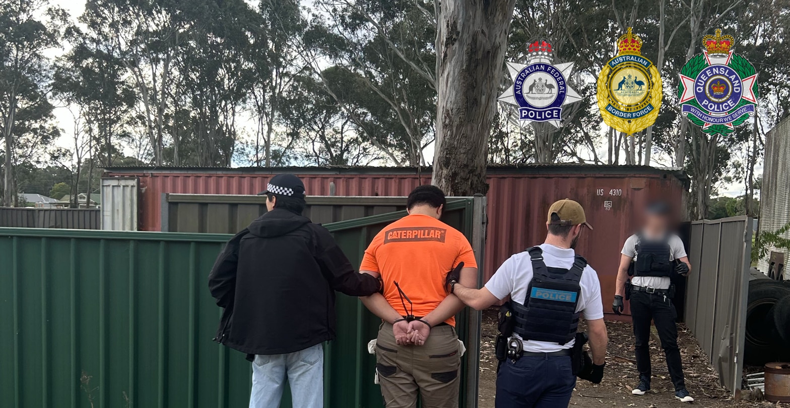 A man in an orange shirt is arrested with his hands behind his back, by three officers. A shipping container is in the backgroun