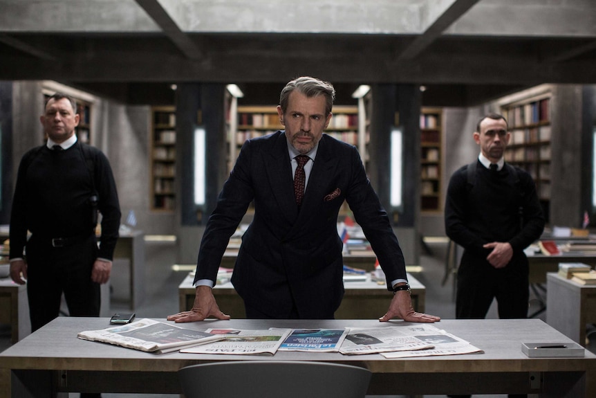 A scene from the movie The Translators with a man in a suit at a desk, two security guards surround him