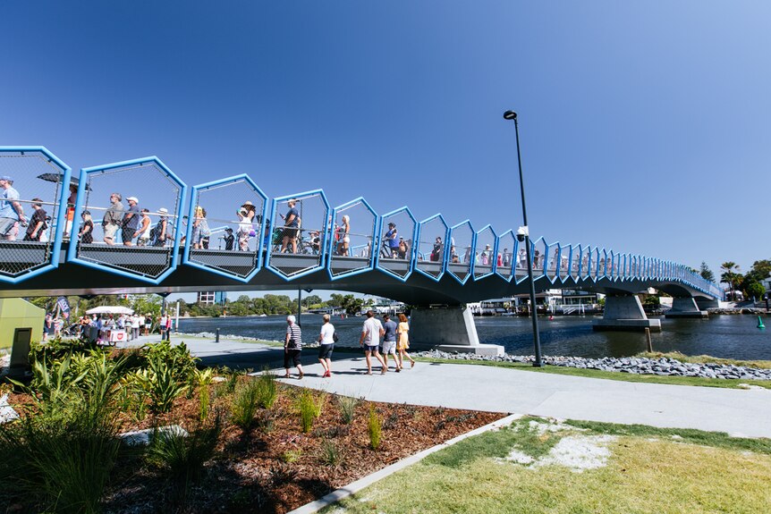 a blue bridge on a sunny day with green plants in the foreground and people walking across it