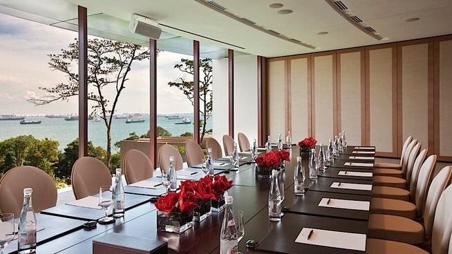 A boardroom table decorated with individual water bottles, glasses, pens and paper. The room has a view of the ocean.