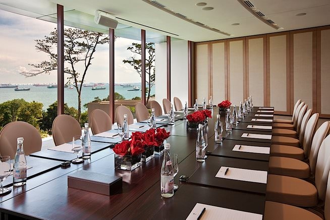 A boardroom table decorated with individual water bottles, glasses, pens and paper. The room has a view of the ocean.