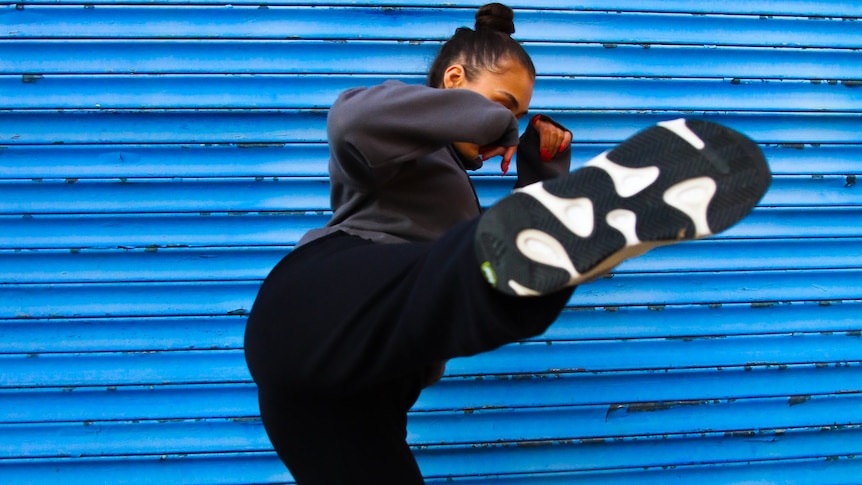 A woman wearing black kicks high into the air against a blue roller door