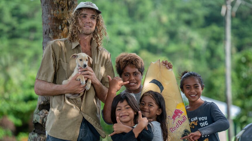 A man in his mid twenties holding a puppy, long curly hair, with some Islander children smiling with a surfboard and big smiles.