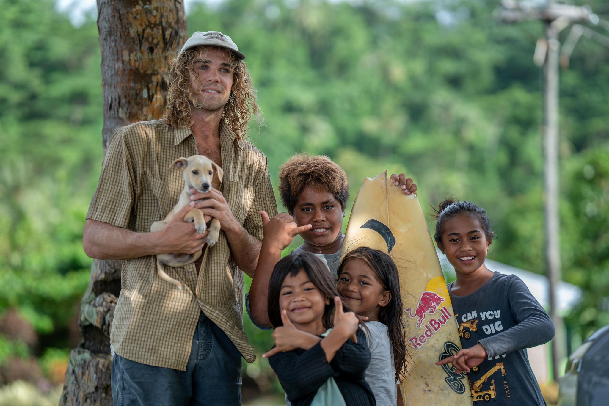 A man in his mid twenties holding a puppy, long curly hair, with some Islander children smiling with a surfboard and big smiles.