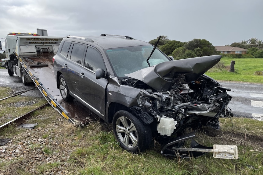 A grey four-wheel-drive with a badly damaged front bonnet is pulled onto a tow truck parked across a train line