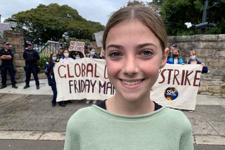 A girl smiles close to the camera as kids hold a banner behind her, while police stand off to the side