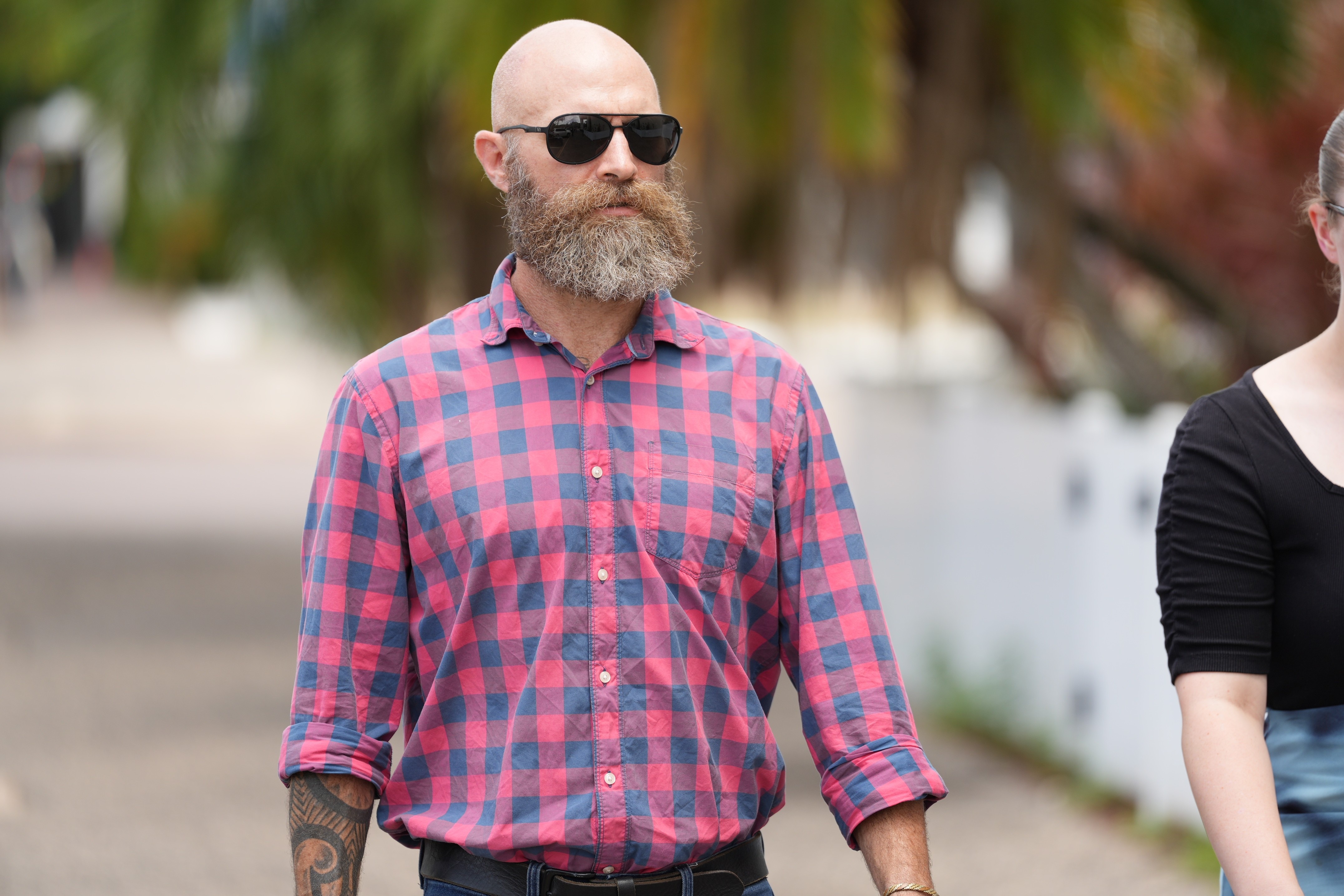 Hombre blanco, calvo, gafas de sol tintadas de negro, barba gris rojiza, vestido con camisa a cuadros rosa y azul