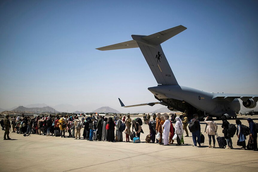 A line of people stand in front of a grey plane while a soldier watches on.