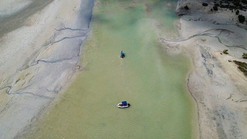 A drone shot of a fishing boat, in a long blue creek with sand on either side, shot from above.