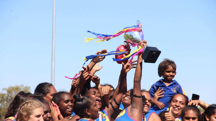 A group of Indigenous women's footballers celebrate a win in their grand final, holding up a trophy.