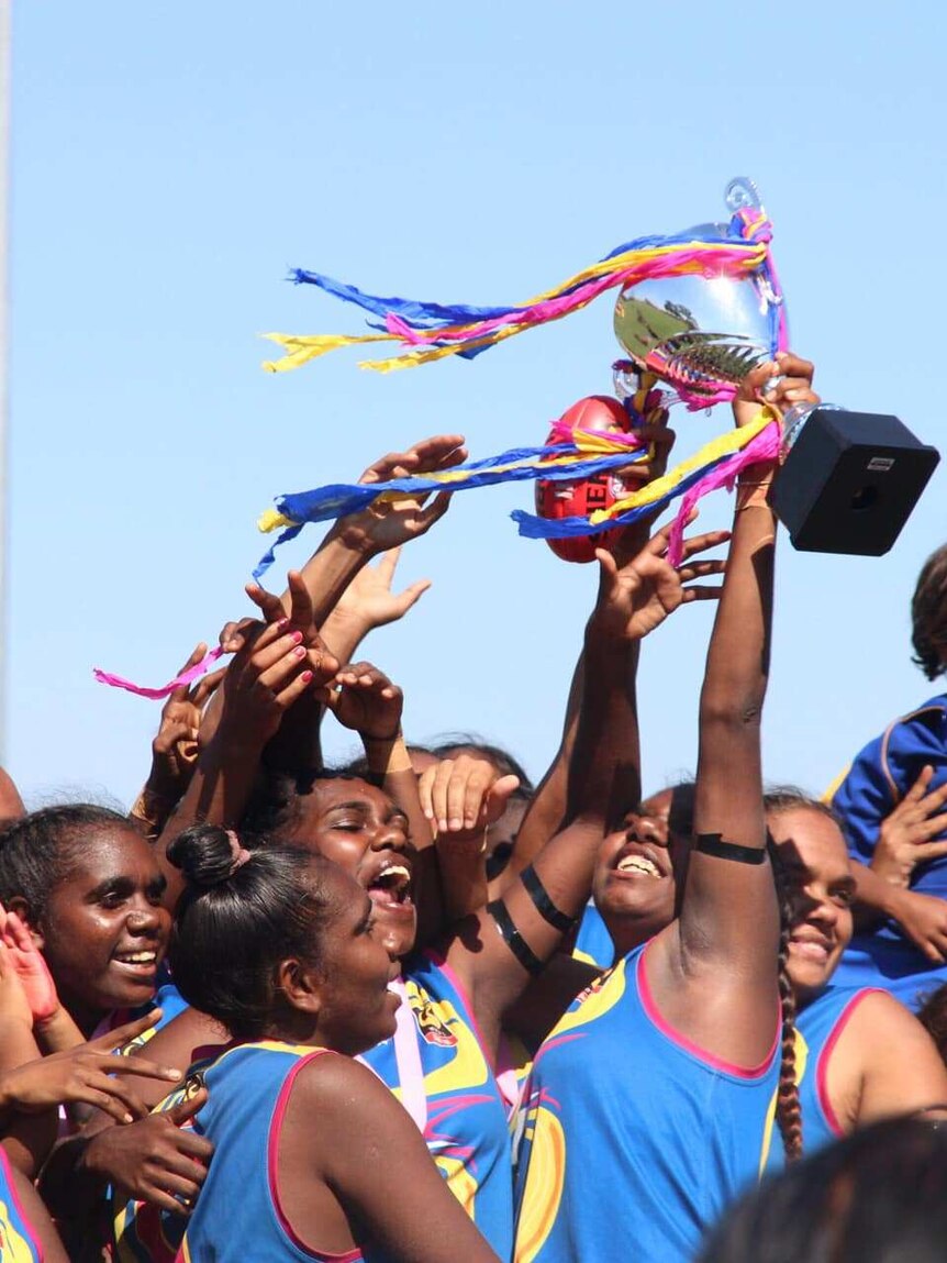 A group of Indigenous women's footballers celebrate a win in their grand final, holding up a trophy.