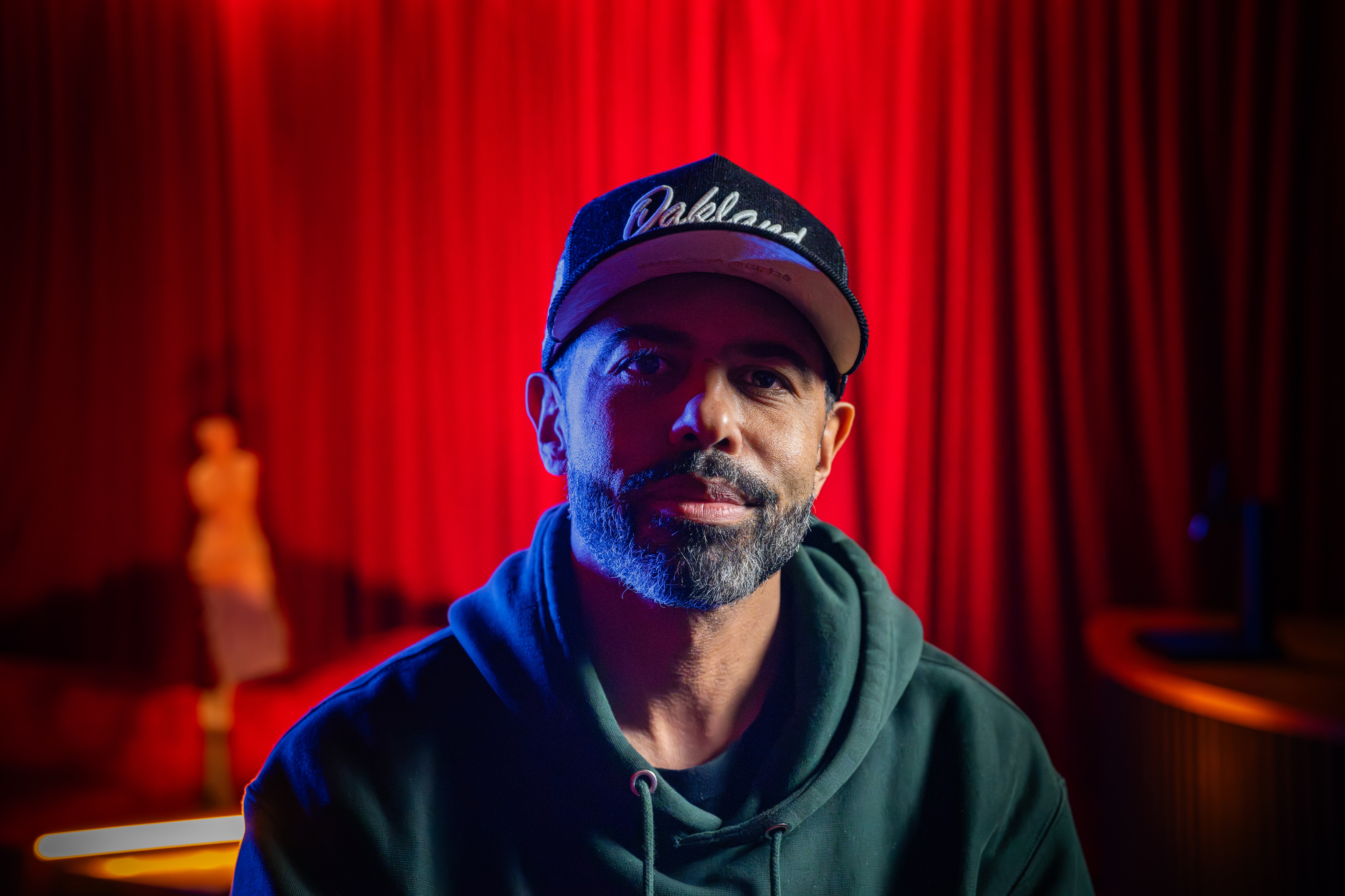 Daveed Diggs sits in front of a red curtain.