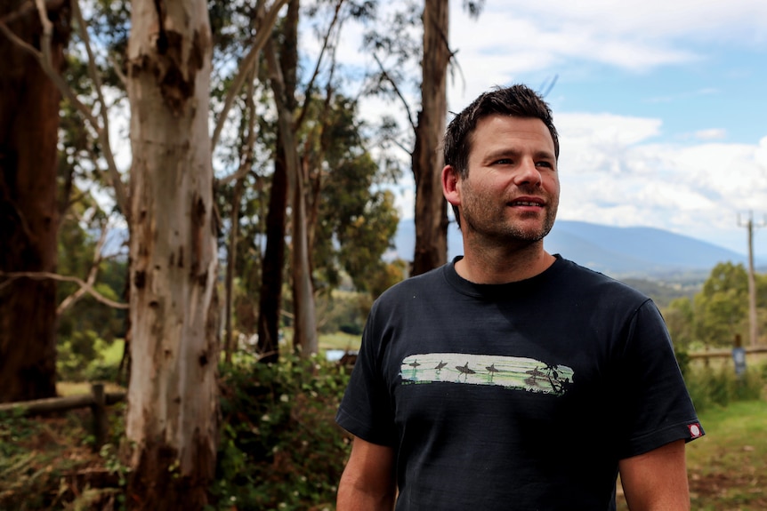 Man with black hair and white skin in blue t-shirt stands in front of trees with mountains in background