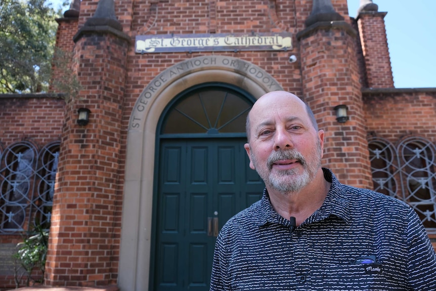 A man wearing a blue and white shirt, stands in front of an old brick church, with the sign St George's Cathedral over the door.