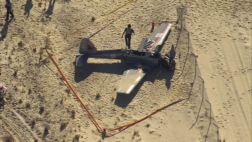 A crashed plane with a damaged nose is on sand and taped off as an emergency services work nearby.