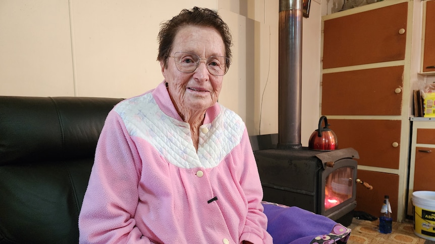 An older woman sits in her home, near a fireplace, wearing a dressing gown.