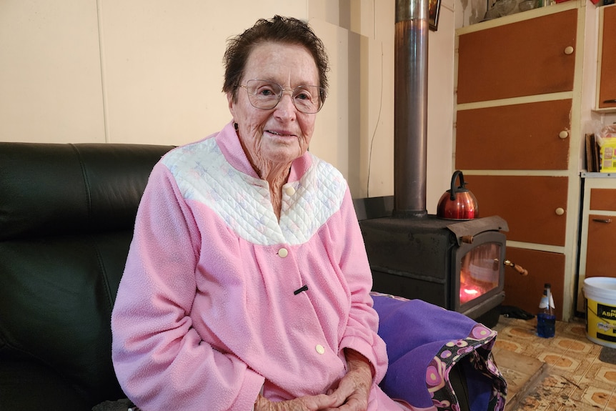 An older woman sits in her home, near a fireplace, wearing a dressing gown.