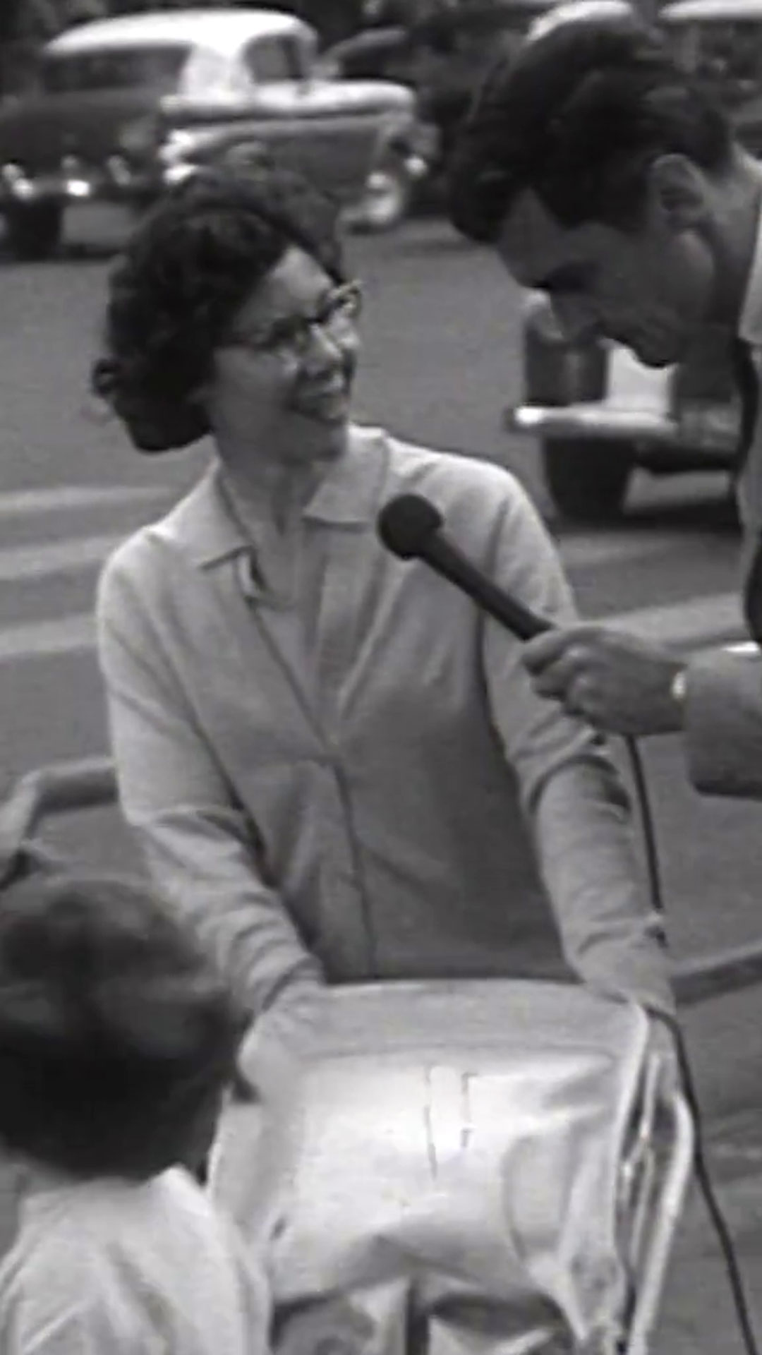 A black and white photo shows a young woman wearing glasses and pushing a pram talking to a man with a mic