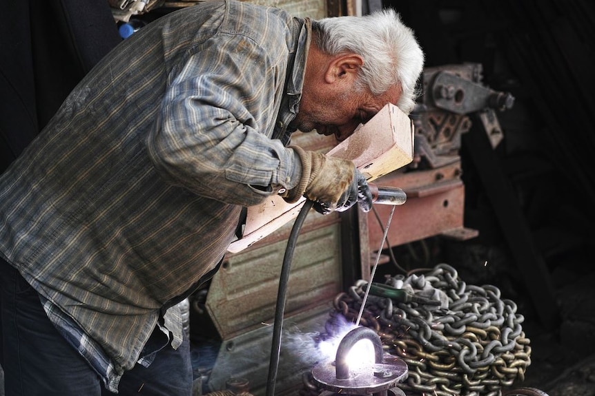 A man with grey hair bends over metal work in a workshop