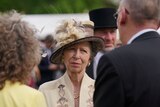 An image of an older lady frowning, she wears a fascinator and looks to be talking to people.