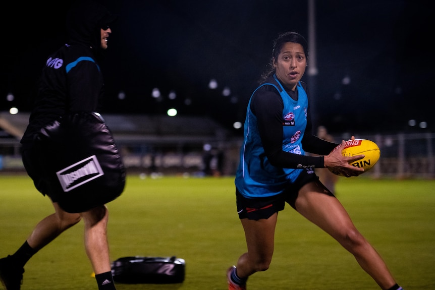 A woman in blue vest playing footy and another person holding a punching bag.