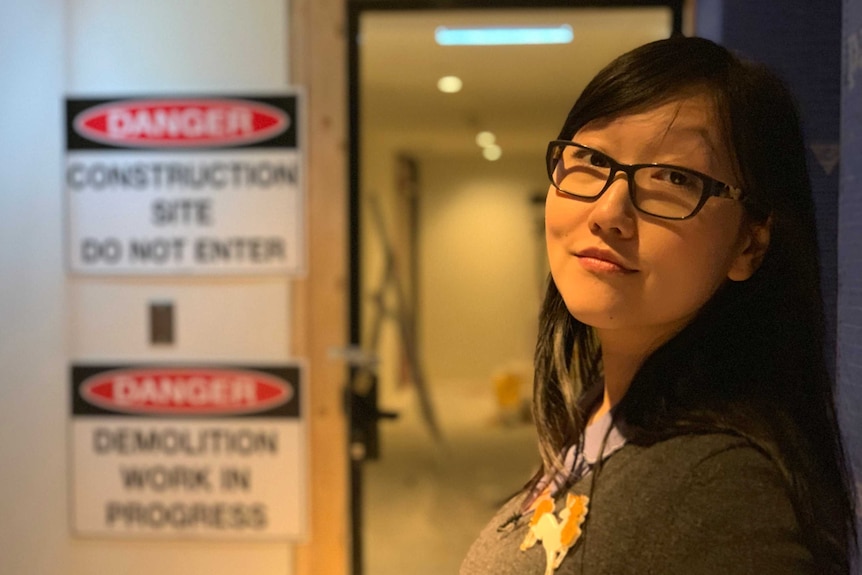 Jenny Zhang stands in front of a doorway with 'construction site do not enter' signs next to it.