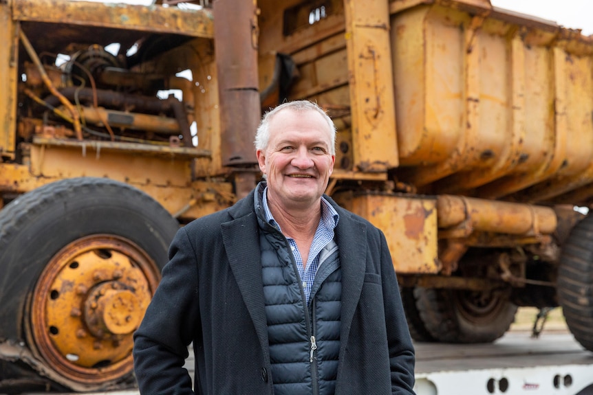 Grey-haired man wearing a black puffer jacket stands in front of an old yellow truck smiling.