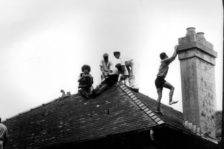 Police and protesters on the roof of a house, with the police detaining one man as another climbs a chimney.