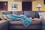 Sick person lies on couch underneath blanket.