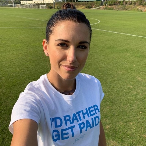 Alex Morgan on a football field taking a selfie and wearing a white T-shirt that says "I'd rather get paid".