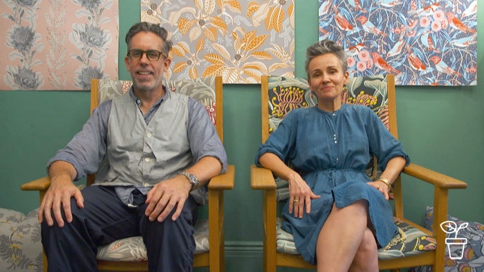 Man and woman sitting on seats with colourful framed fabrics on wall behind them