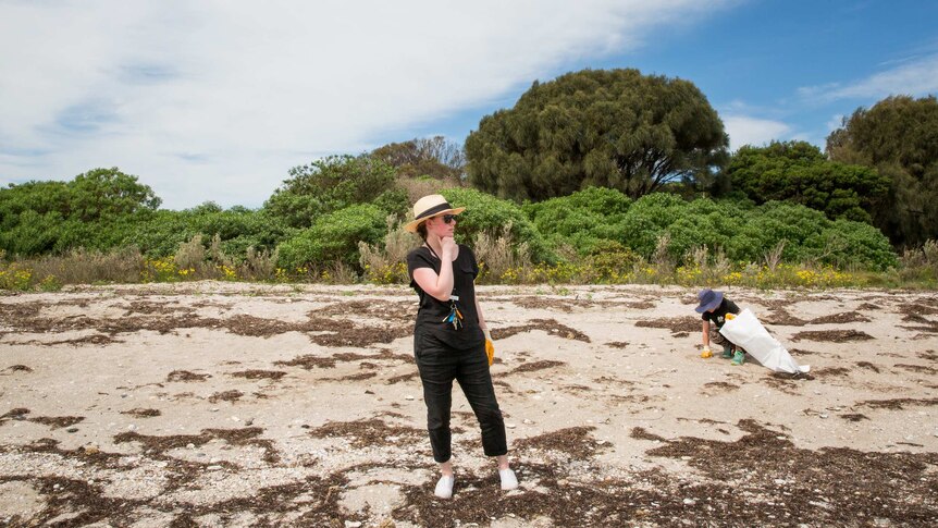 Teacher Anita Harding, wearing hat and sunglasses, surveys the beach while a student crouches, putting marine debris into a bag.