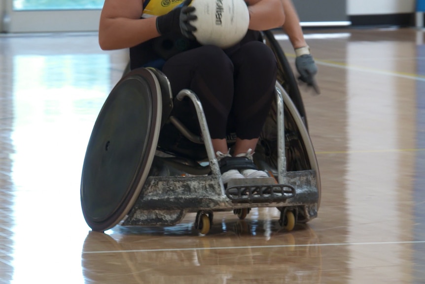 An unidentifiable person in a wheelchair rugby chair with a reinforced front bumper, holds a white ball.