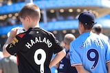 Two young Manchester City fans, one drinking from a bottle, in Erling Haaland jerseys with other fans outside the stadium