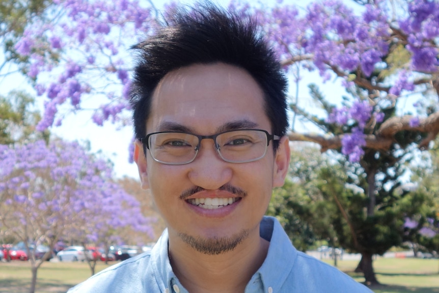 A headshot of a man with black hair and glasses smiling with purple jacaranda trees in the background.
