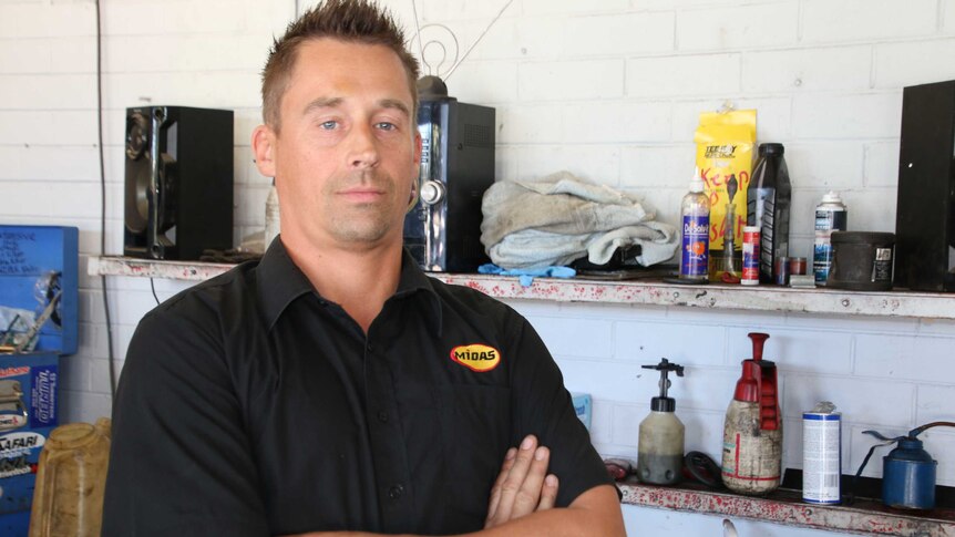 A man in a black workshop uniform looks at the camera with a serious expression in a workshop.