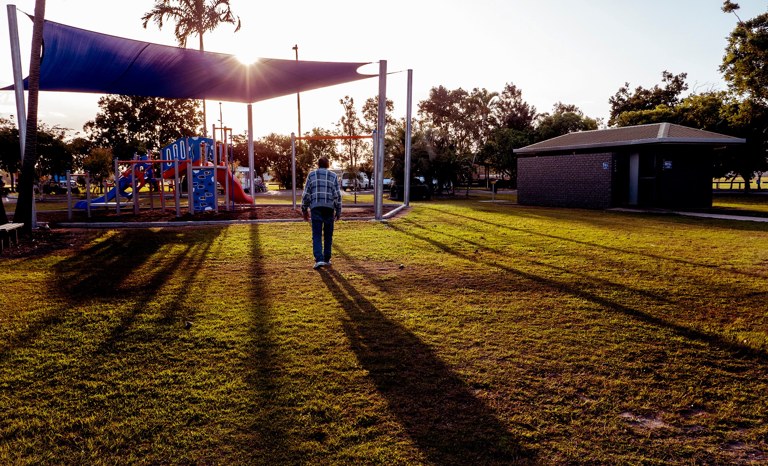 A playground and toilet block at sunset as a man walks with back to camera on grassy land.