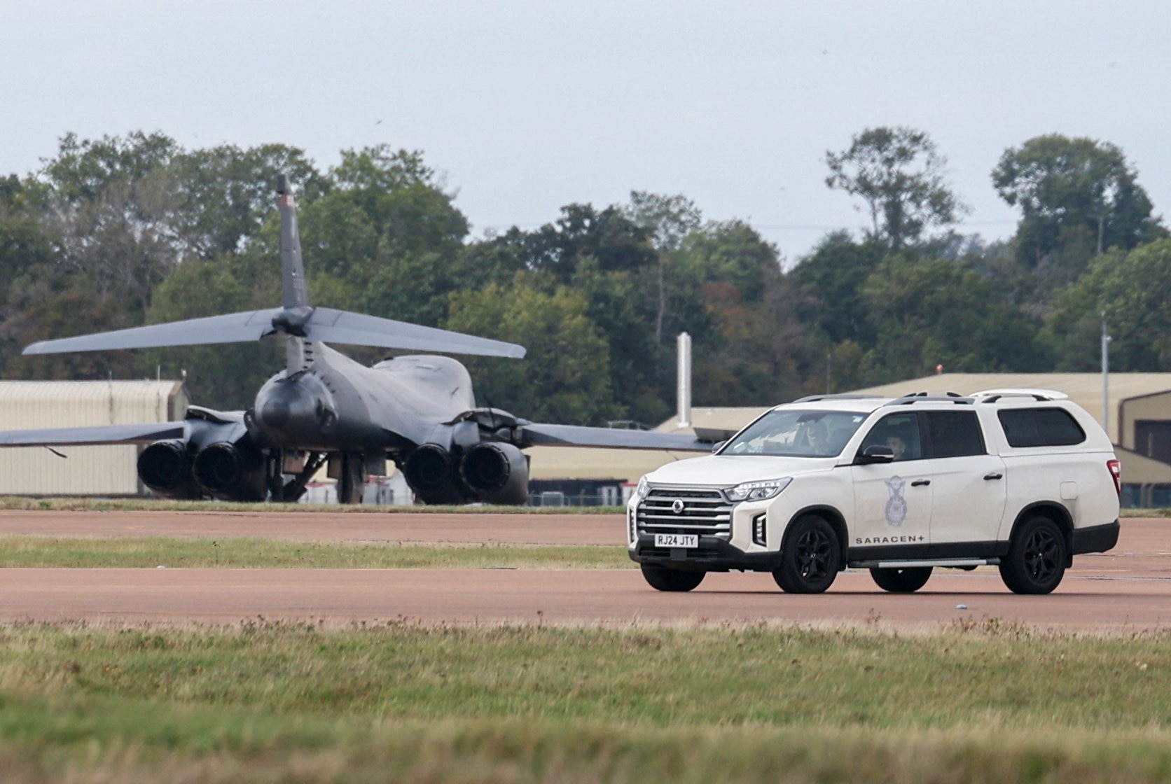 A large, dark grey military plane seen on a grass and dirt runway near to a white police SUV.