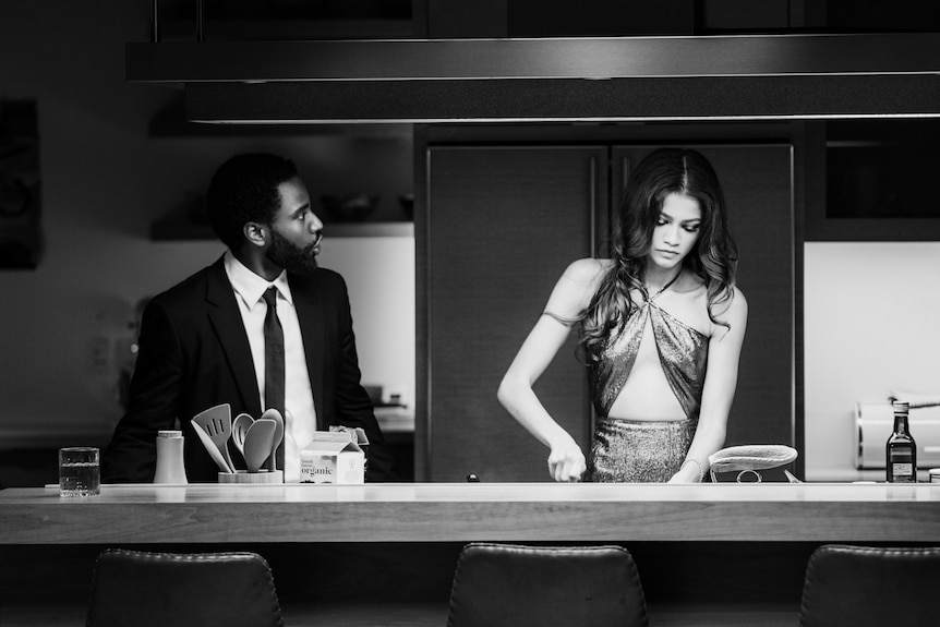 Black and white photo of young man in suit and woman in dress, standing at kitchen bench as if talking while she cuts something.