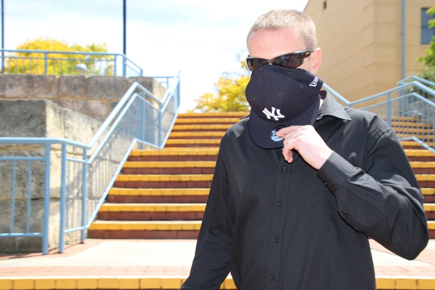 Luke Kevin Dempster covers his face with a cap as he leaves court. He is wearing a black shirt and has on sunglasses.