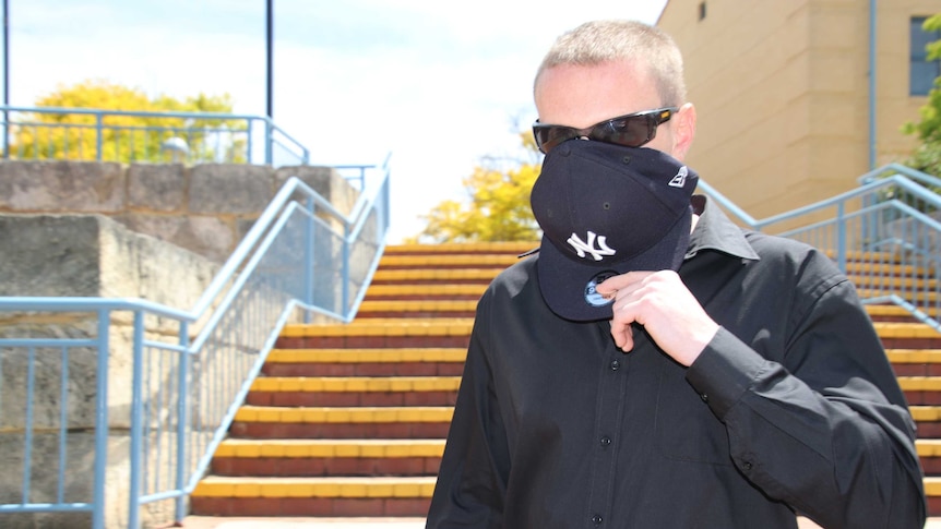 Luke Kevin Dempster covers his face with a cap as he leaves court. He is wearing a black shirt and has on sunglasses.