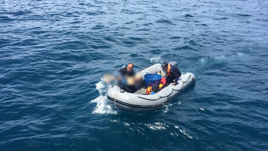A rubber raft in the middle of a blue sea with two men on board, one carrying a man's body.