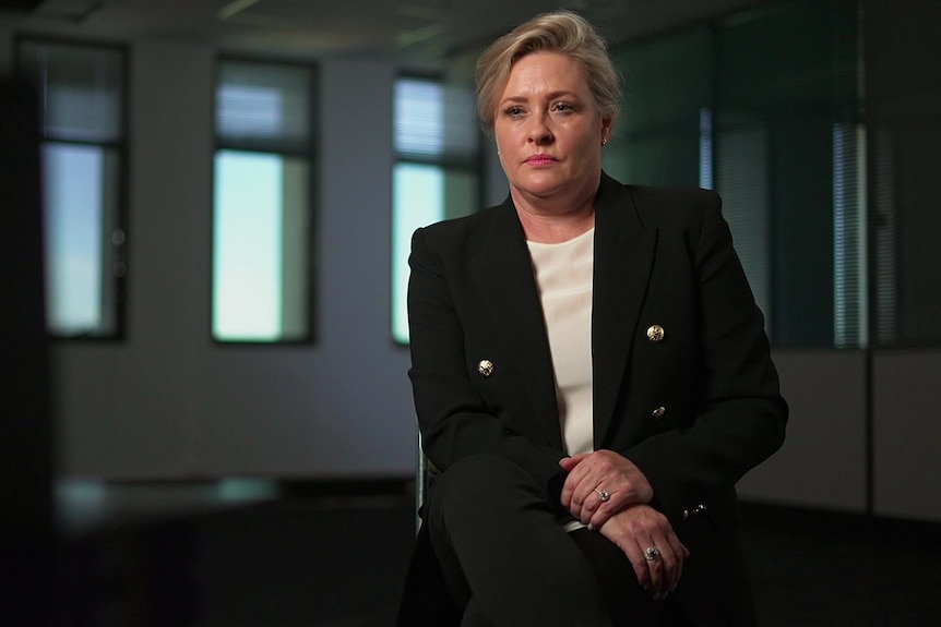 A woman is seated wearing a dark suit jacket over a white top.