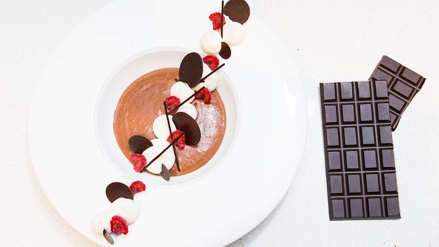 Chocolate mousse with raspberry and chocolate garnish in a white bowl, next to a dark chocolate bar.