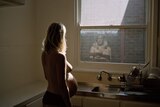 Portrait of heavily pregnant woman in kitchen, back to camera, looking downwards to kitchen sink as light falls through window.