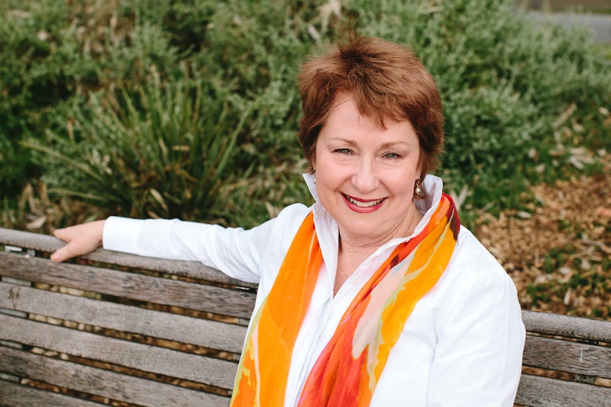 A smiling, middle-aged woman with short hair, wearing a bright scarf, sits on a park bench.