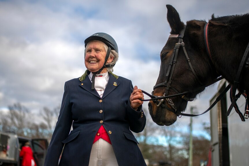 An older woman in a navy blazer and black helmet grins next to her large, muscular, chocolate-coloured horse.