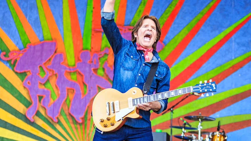 John Fogerty plays guitar on stage before a colourful backdrop. He wears a blue denim shirt.