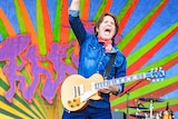 A man in a denim shirt holds his arm aloft while performing with a guitar in front of a colourful backdrop.
